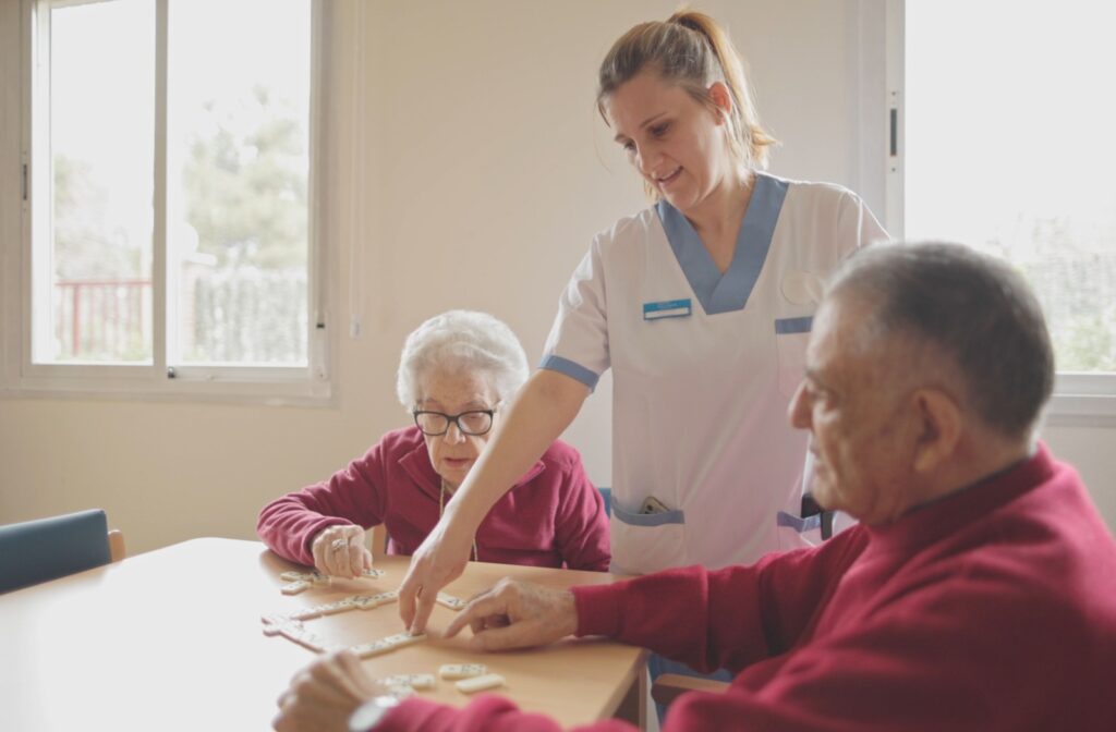 Nurse in white and blue scrubs helping seniors play dominoes on a table.