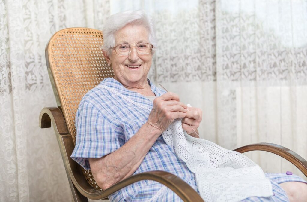 Elderly woman in a wicker chair crotcheting a white doily and smiling.