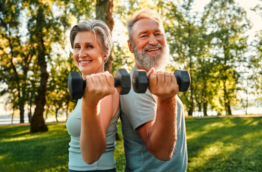 A senior couple each holds a five-pound weight while outside on a warm, sunny afternoon as part of physical fitness