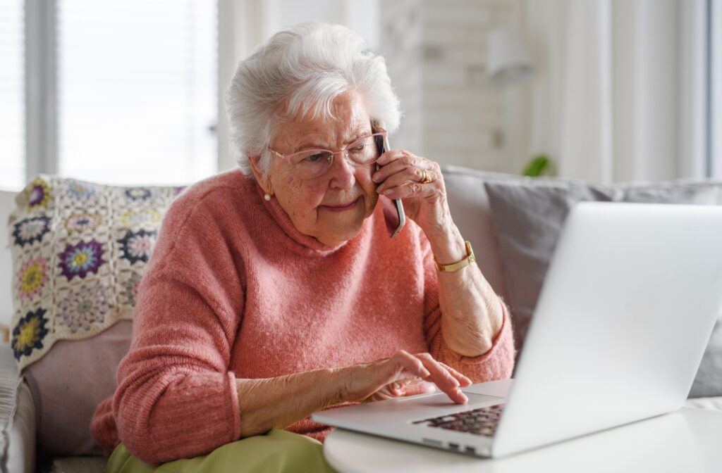 A senior works on a laptop, looking at assisted living communities, while making a call to schedule a tour.