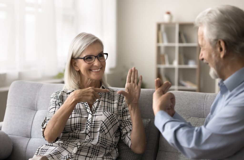 A smiling senior in glasses with a hearing disorder uses hand gestures, showing symbols, to communicate with another senior.