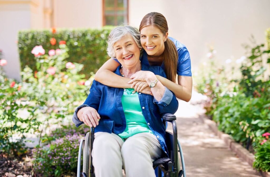 A caregiver hugs a seated resident from behind while they take a walk together through a flower garden in the courtyard of their community
