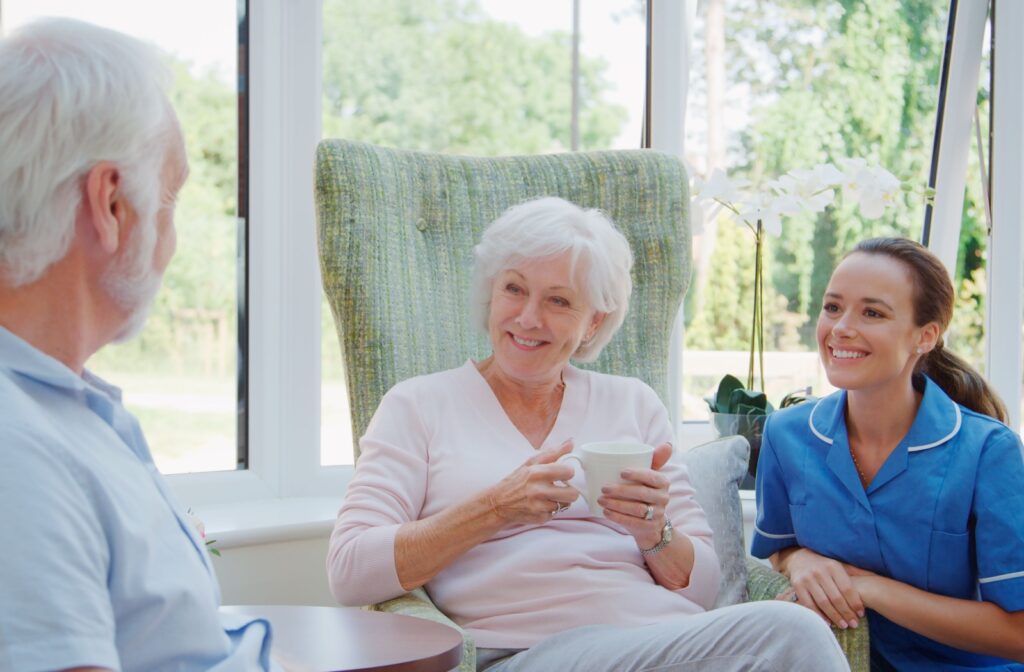 Two residents in assisted living sit in front of a bright window and enjoy a cup of tea over conversation while a caregiver kneels to check in on them