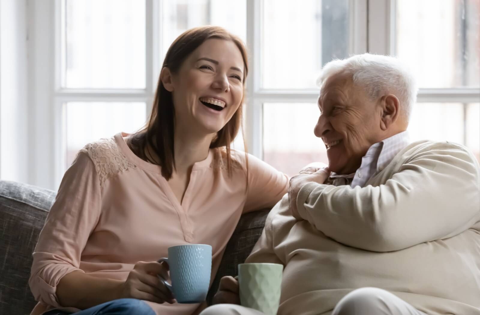 A senior and their younger loved one laugh together over a cup of coffee